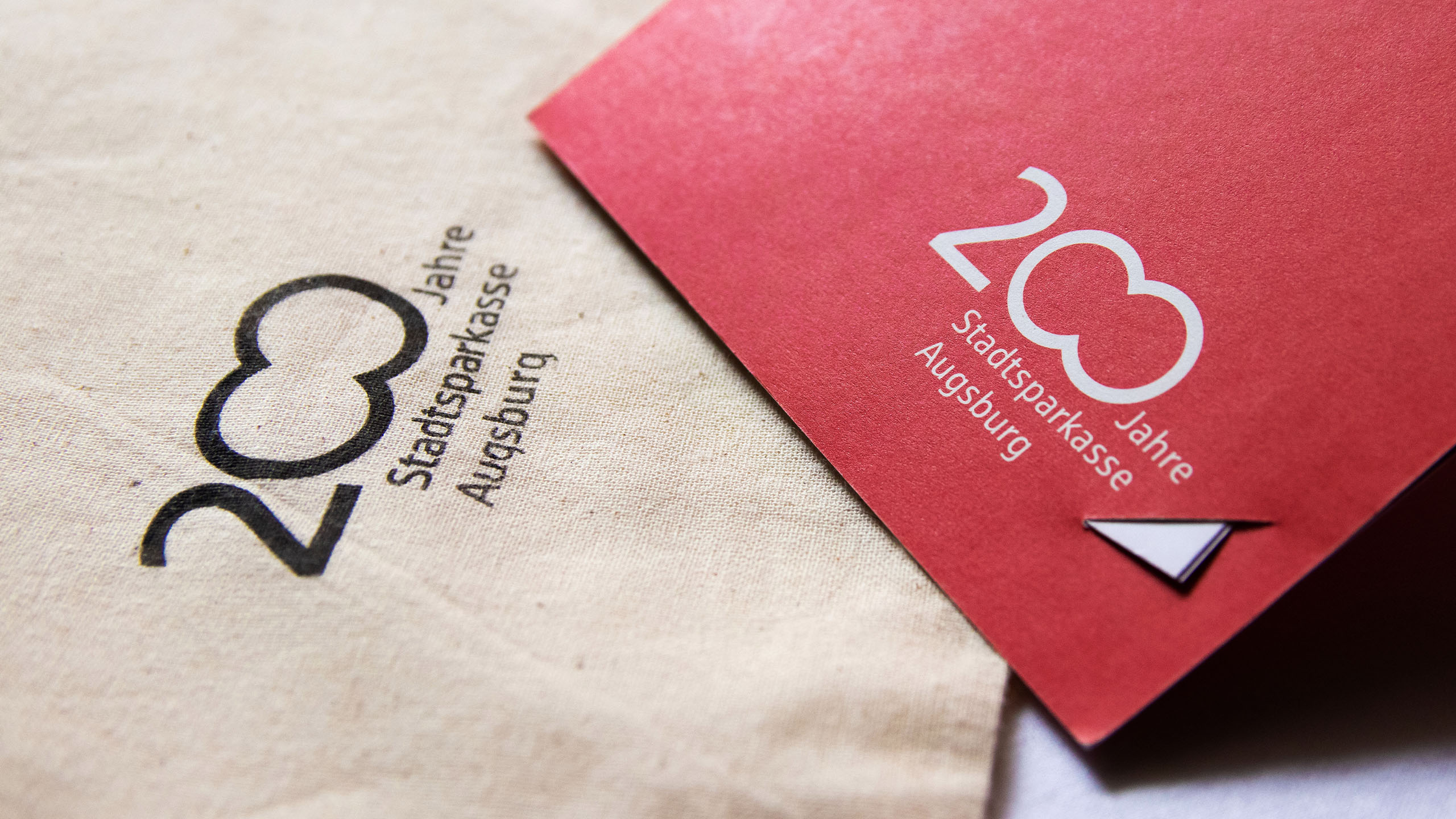 Das 200-Jahre-Logo der Stadtspaarkasse Augsburg auf einem Jutesack und einem roten Karton. BRANDSTAGE Design